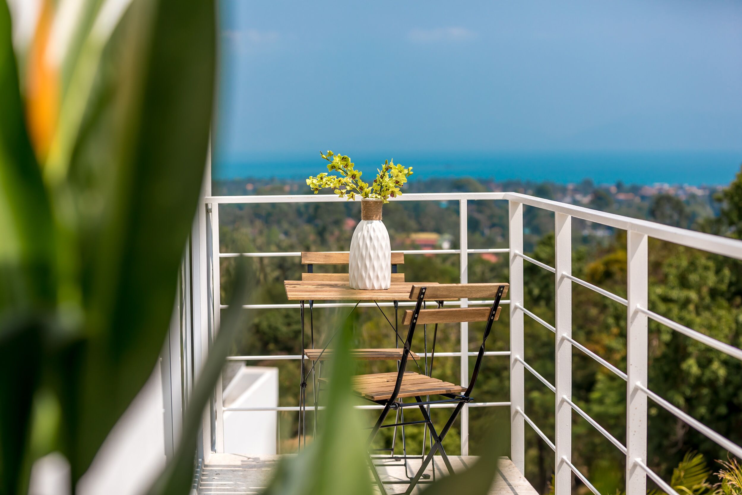 A balcony with a white railing, a wooden table, and a white vase with yellow flowers.