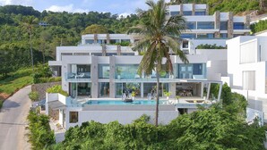 Une maison moderne à plusieurs étages, dotée d’une piscine, entourée d’une végétation luxuriante et située au pied d’une colline.