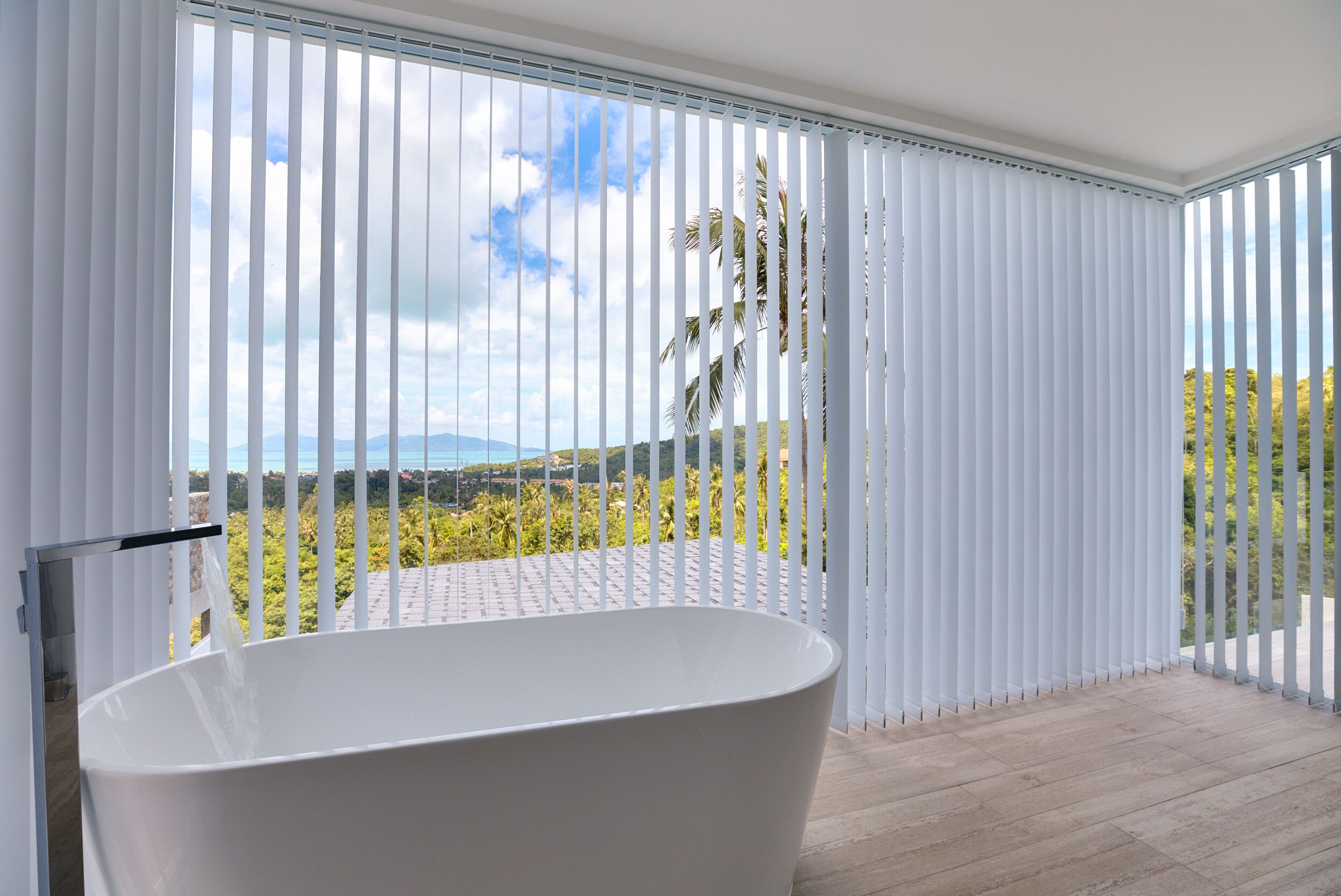 A modern bathroom with a freestanding bathtub, large windows with vertical blinds, and a view of a mountainous landscape.
