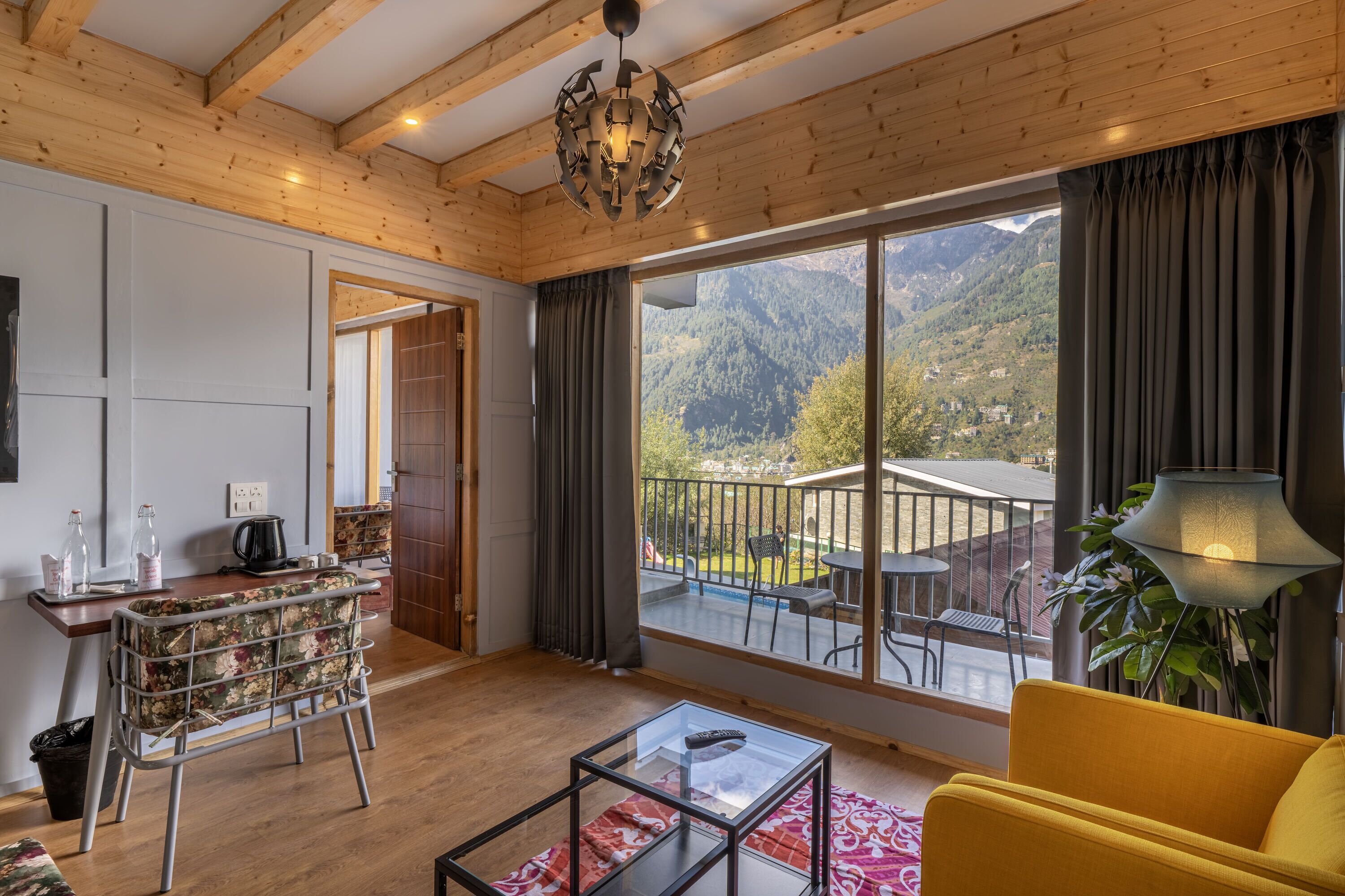 A room with a wooden ceiling, a large window with curtains, a yellow sofa, a glass coffee table, and a metal chair.