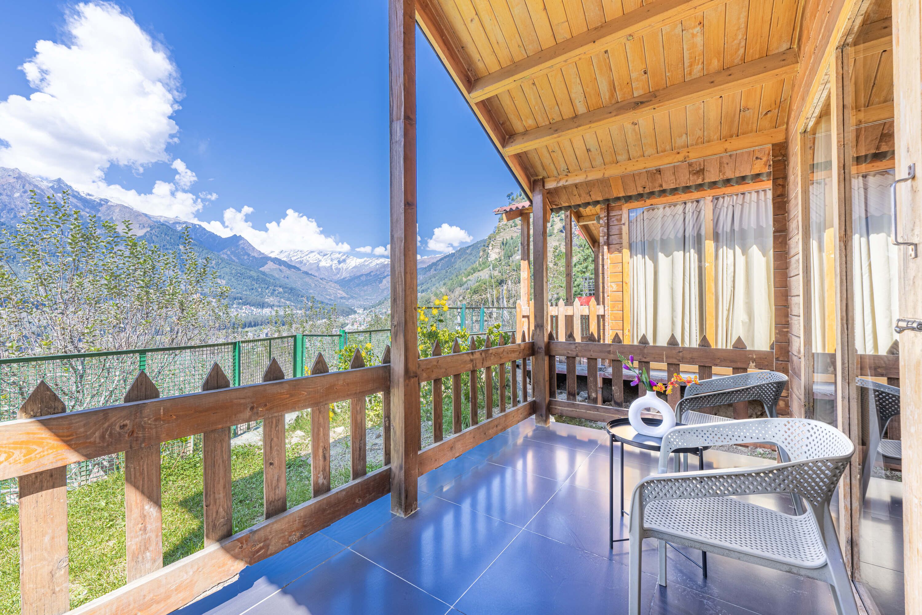 Un balcón de madera con vistas a montañas, una mesa con sillas y una barandilla.