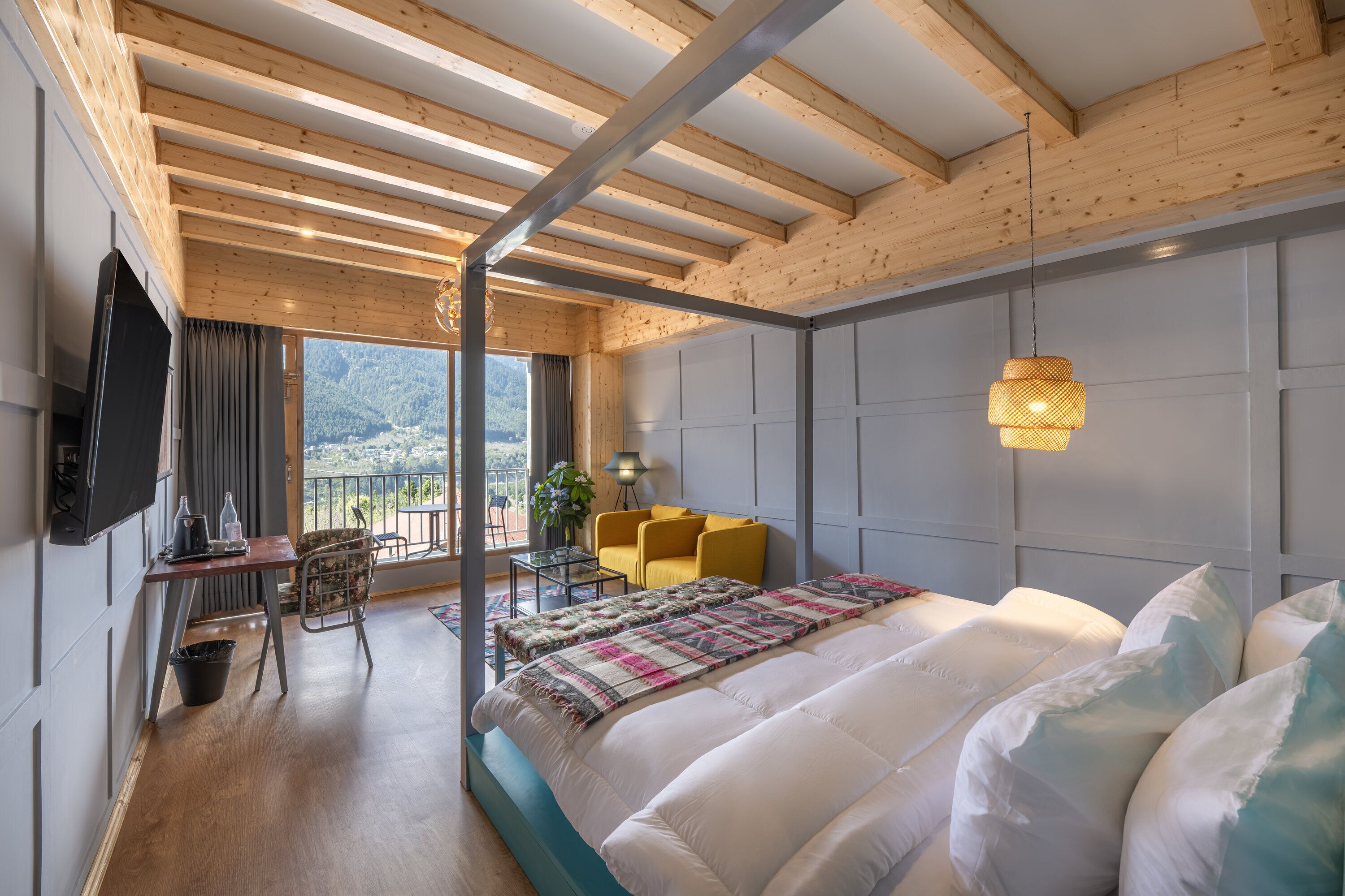 A bedroom with a large bed, a desk, a chair, a TV, and a balcony with a view of the mountains.