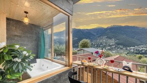 A balcony with a view of mountains, a wooden deck, a black chair, a dining table with food and drinks, and a spa area with a bathtub and a plant. - Echor Himalayan Heights Luxe Resort & Spa, Manali (Manali)