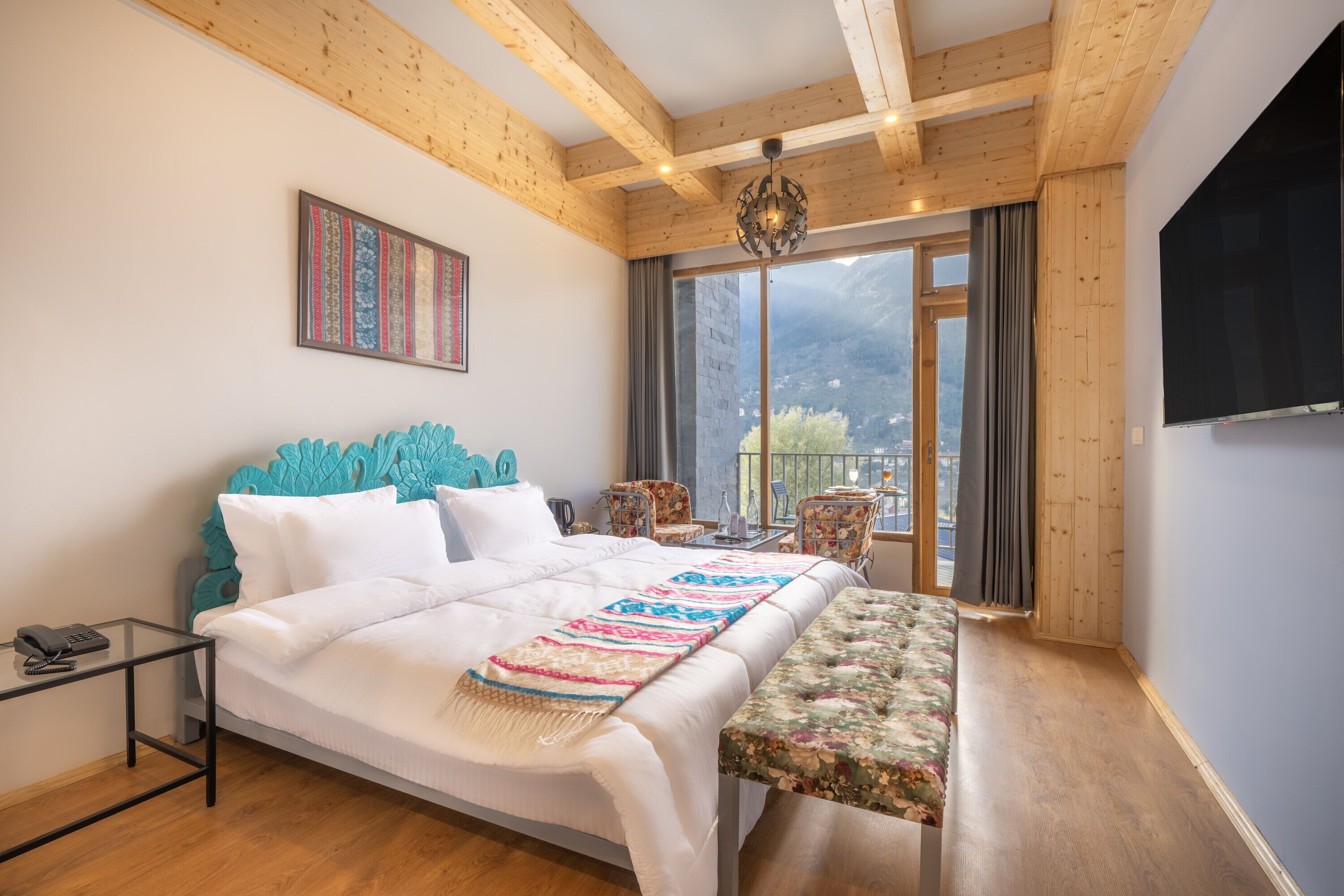 A bedroom with a wooden ceiling, a bed with a white headboard, a bench, a small table, and a TV mounted on the wall.