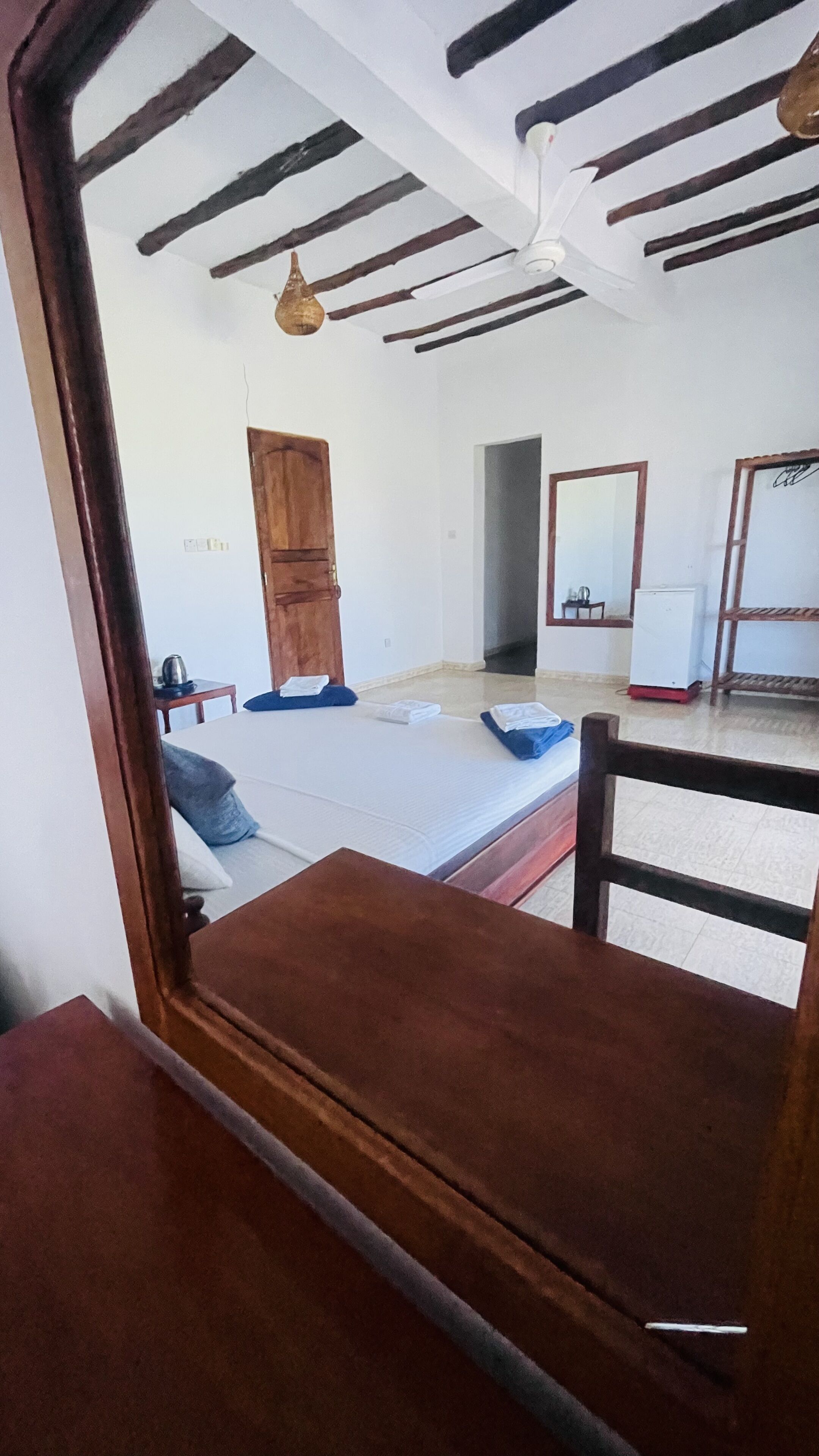 A room with a bed, a wooden table, a ceiling fan, a wardrobe, and a mirror.