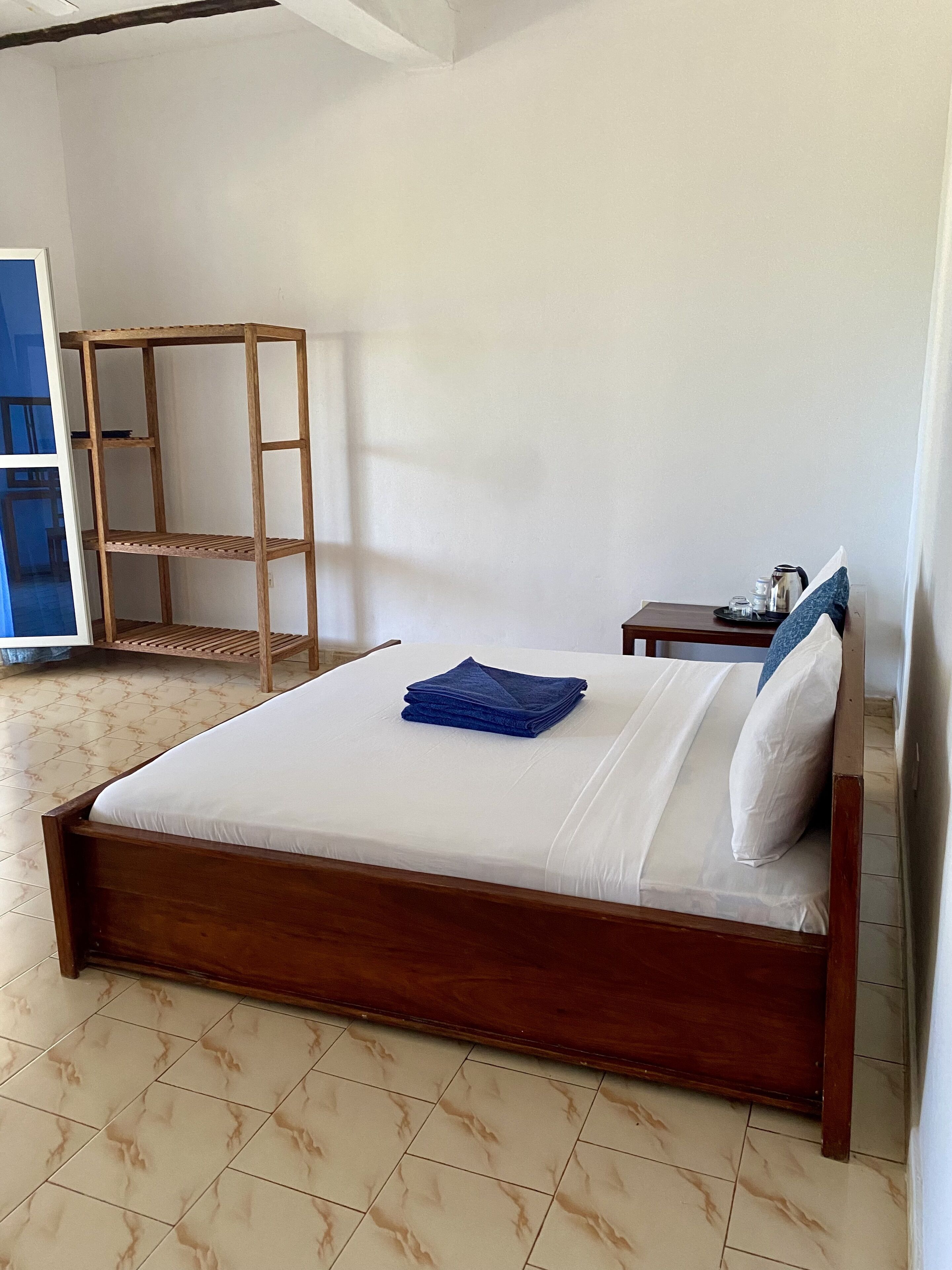 A neatly made bed with a blue towel, a wooden nightstand with a kettle, and a wooden shelf unit in a room with tiled flooring.