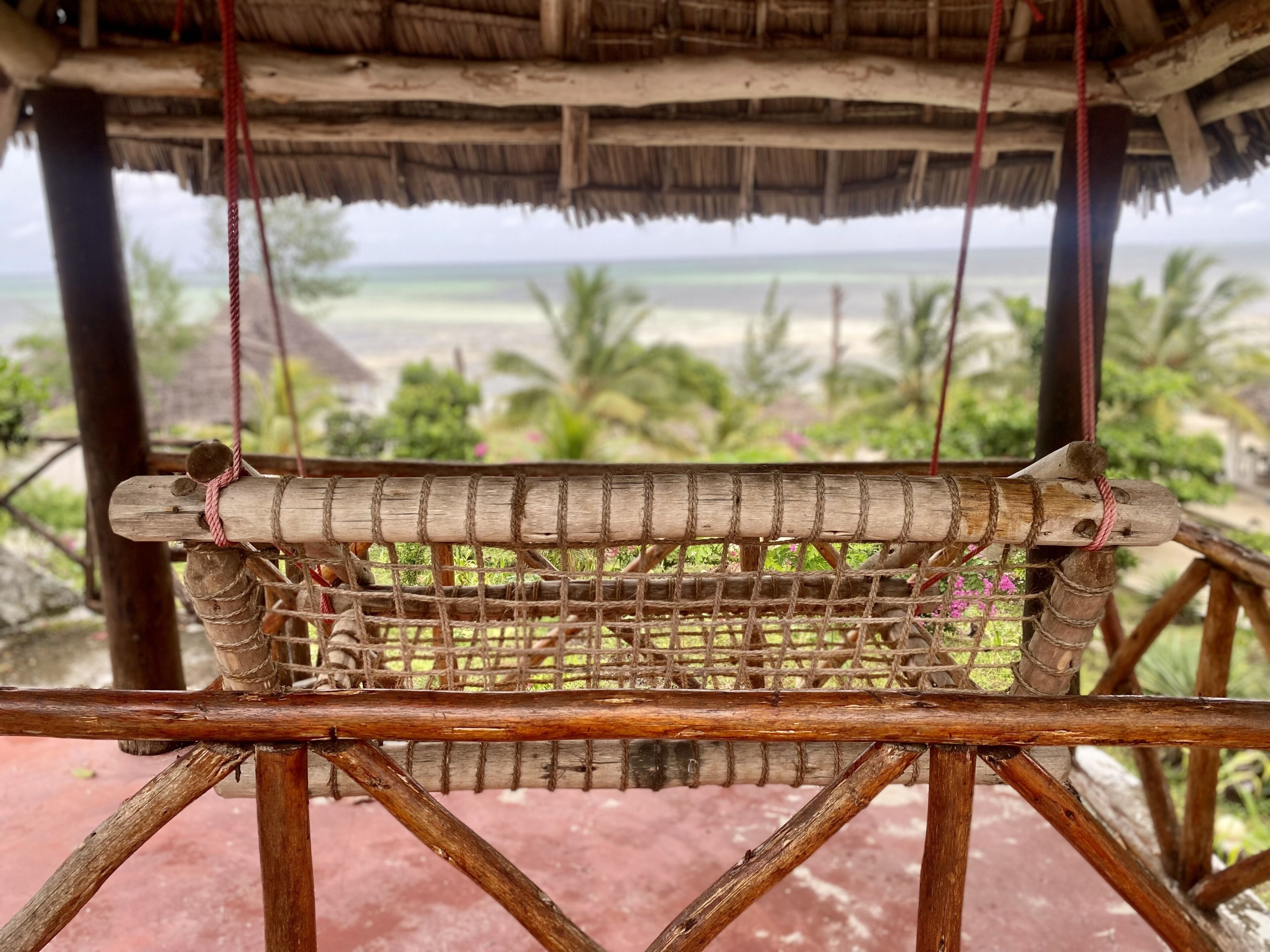 A hammock suspended between wooden beams with a view of a beach and palm trees in the background.