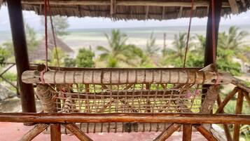 A hammock suspended between wooden beams with a view of a beach and palm trees in the background.