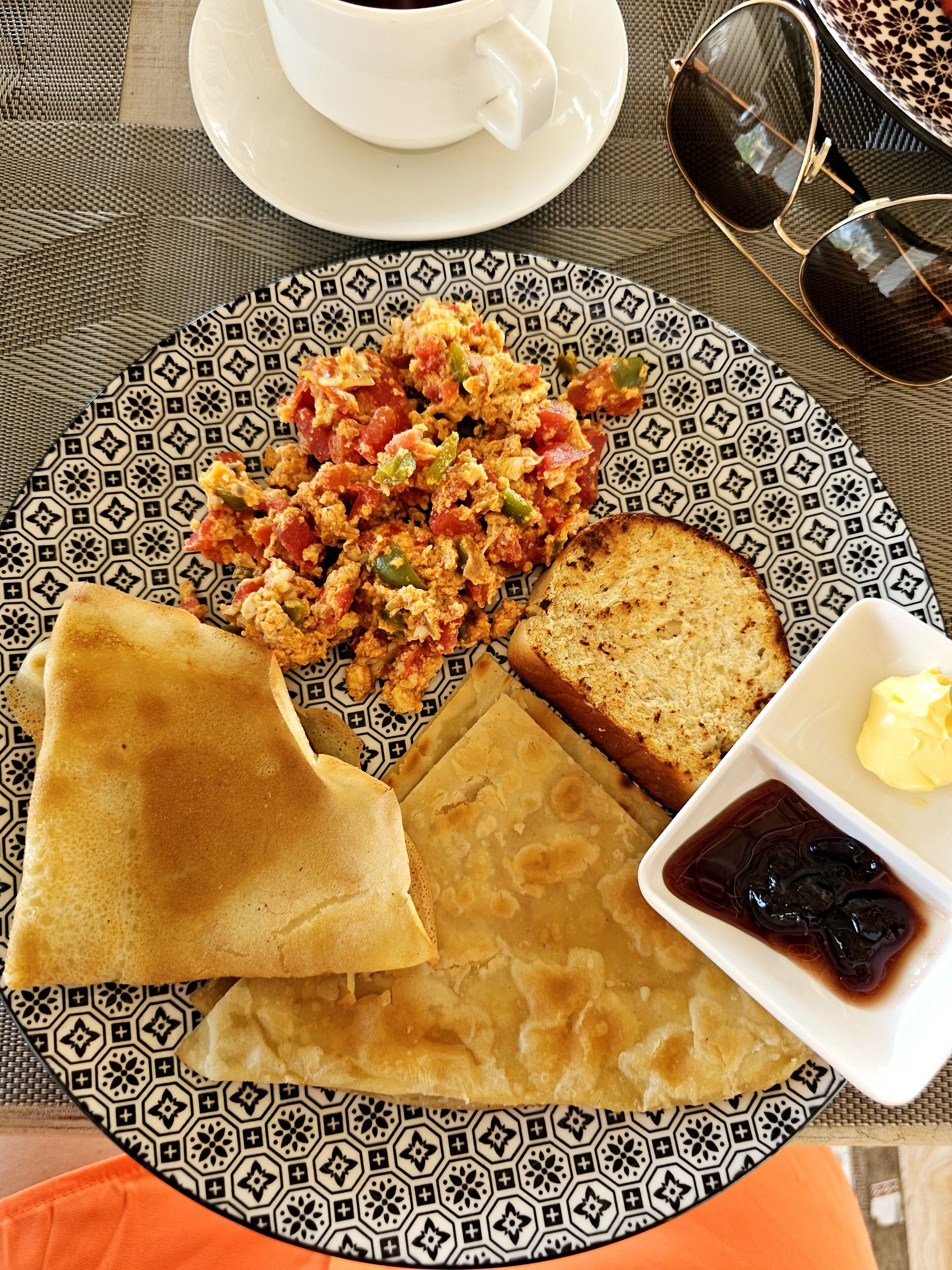 Un plato de comida con huevos revueltos, tostadas y una rebanada de pan, acompañado de un recipiente pequeño de mermelada y una taza de café sobre un mantel estampado.