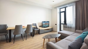 Een moderne woonkamer met een bank, een eettafel met stoelen, een televisie en een groot raam met gordijnen.