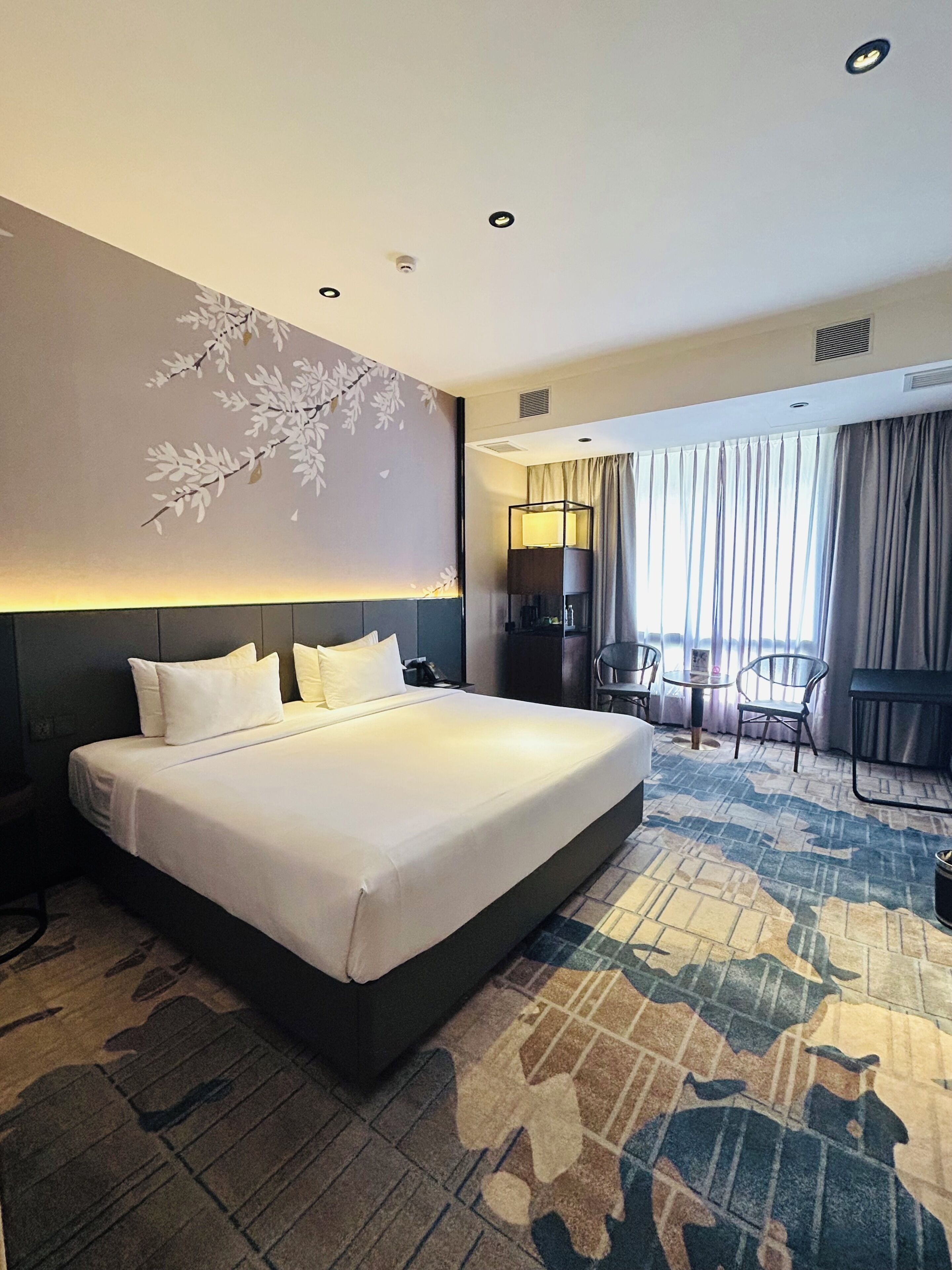 A hotel room with a large bed, a desk with a chair, a lamp, and a view of the outside through a window with curtains.