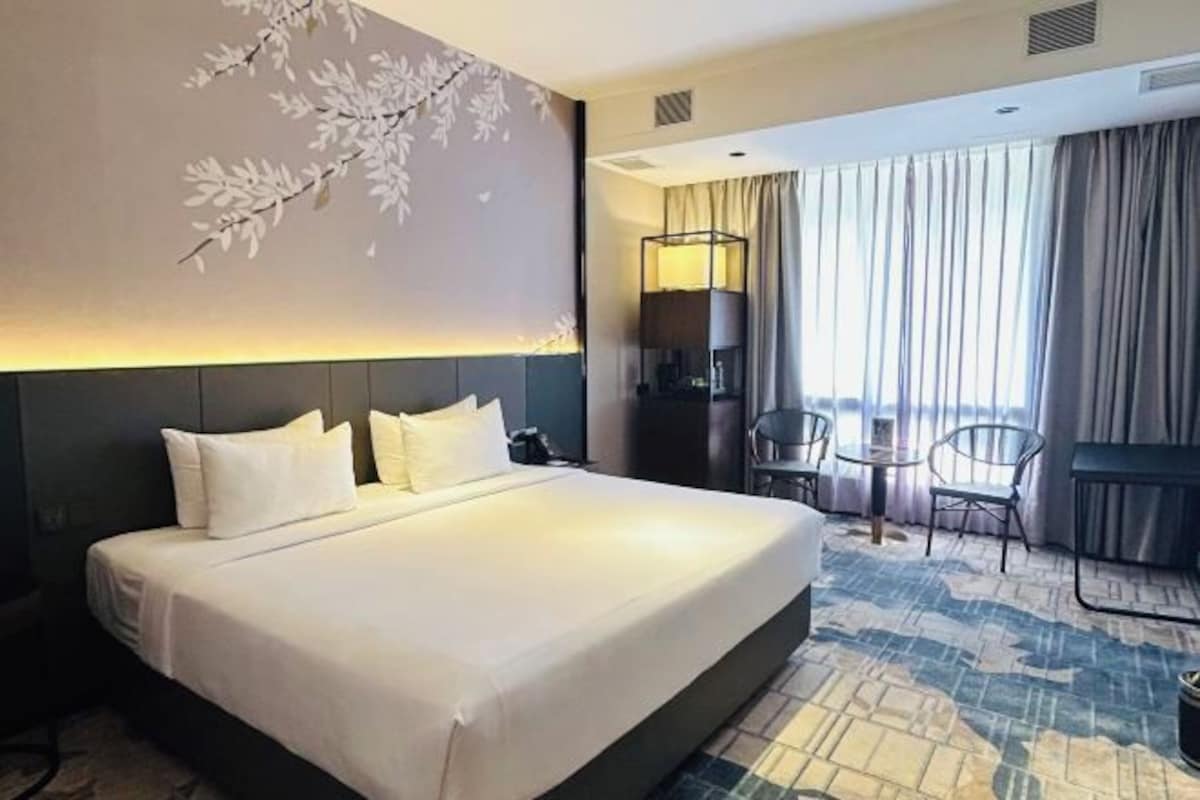 Habitación de hotel con una cama grande, un escritorio y una silla. Se ve la ciudad a través de la ventana.