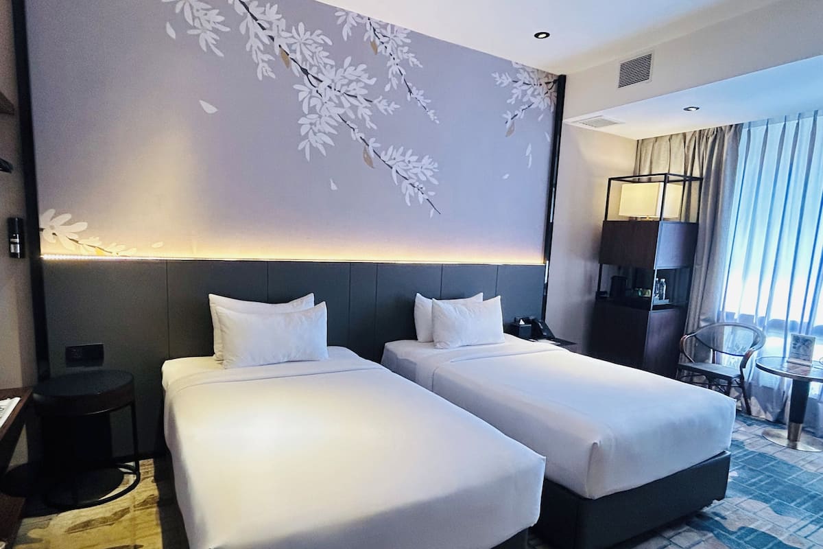 A hotel room with two beds, a desk, a chair, a lamp, and a large window.