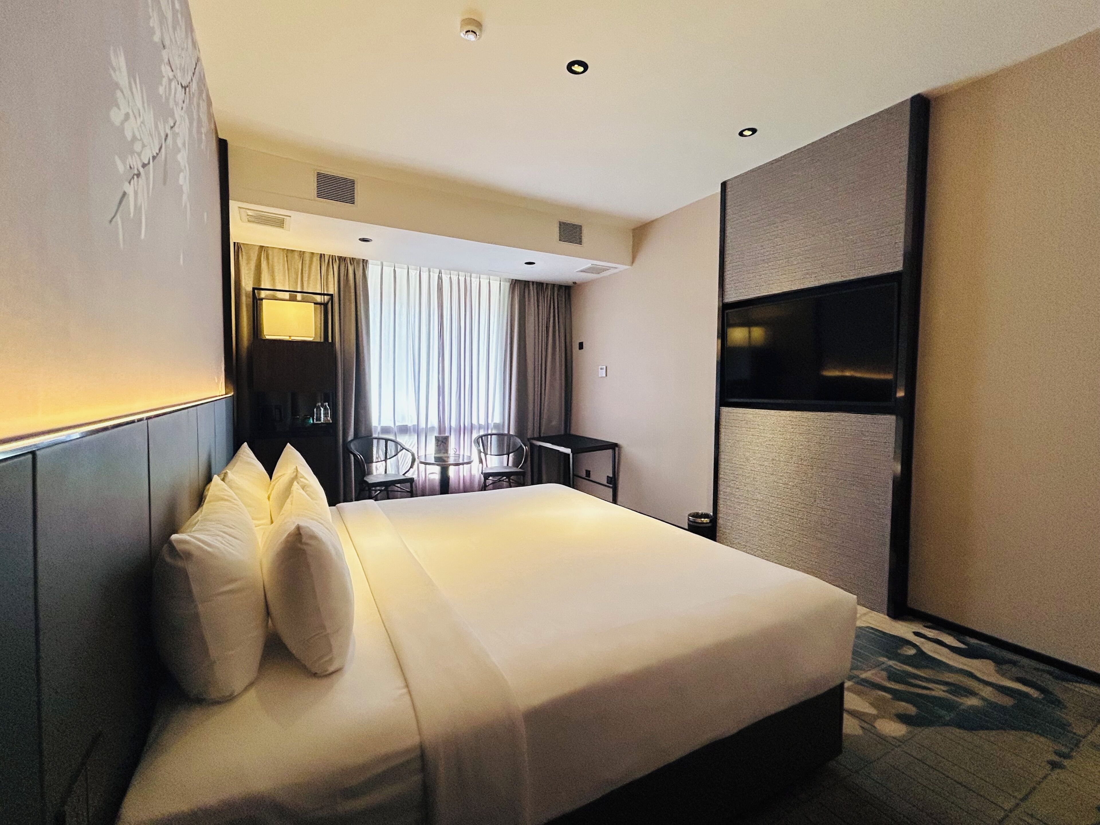 A hotel room with a large bed, a desk, two chairs, a TV, and a window with curtains.