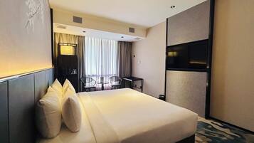 A hotel room with a large bed, a desk, two chairs, a TV, and a window with curtains.