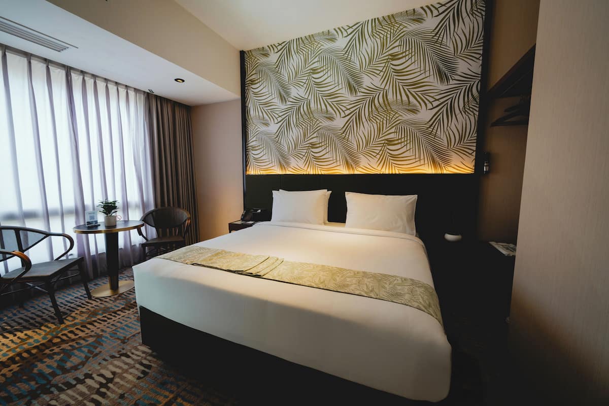 A hotel room with a large bed, a headboard with a leaf pattern, a small table with a plant, and a chair.