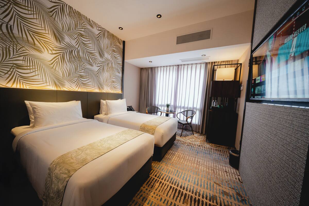 A hotel room with two beds, a patterned wall, a large window with curtains, a small table, and a TV.
