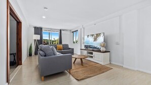 A modern living room with a grey sofa, a round coffee table, and a flat-screen TV on a white cabinet. - Seaside Rest at Clifton Beach (Clifton Beach)