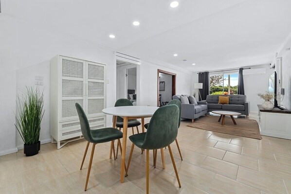 A modern living room with a dining area, grey sofas, a white cabinet, and a large window. - Seaside Rest | 5 Bedroom Beachfront Holiday Home (Clifton Beach)