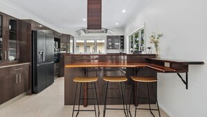 A modern kitchen with dark wood cabinets, a stainless steel refrigerator, a wooden bar counter, and three bar stools. - Seaside Rest at Clifton Beach (Clifton Beach)