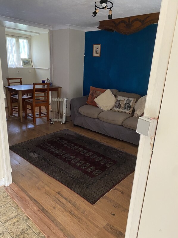 A living room with a blue wall, a grey sofa with patterned cushions, a wooden dining table with chairs, and a dark rug. - Cottage à la ferme (Lengronne)