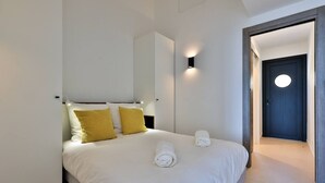 A modern hotel room with a bed, two pillows, a nightstand, a wall-mounted light, and a door with a circular window. - 2-room apartment with panoramic sea view (Rayol-Canadel-sur-Mer)