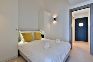 Moderni hotellihuone, jossa on sänky, kaksi tyynyä, yöpöytä, seinään kiinnitetty valaisin ja pyöreäikkunainen ovi.