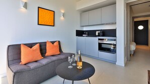 A modern living area with a sofa, a small round table with wine glasses and a bottle, a kitchen with grey cabinets, and a framed picture on the wall. - 2-room apartment with panoramic sea view (Rayol-Canadel-sur-Mer)