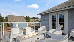 Una terraza con mobiliario exterior blanco, un brasero y un ventanal corredero que da a un área cubierta.