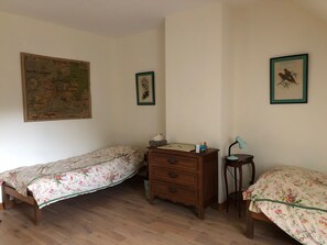 A small, simple room with two single beds, a wooden dresser, a desk with a lamp, and a map on the wall.