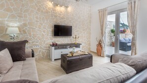 Ett vardagsrum med en vägg av sten, en platt-TV, ett vitt konsolbord, en brun lädersoffa, en läderfåtölj, en byrå i trä och en skjutdörr i glas som leder ut till ett utomhusområde med växter och utsikt över en byggnad.