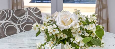 A white table with a vase of white flowers, curtains, and a view of a pool.