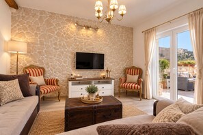 Una sala de estar con una pared de piedra, un televisor de pantalla plana, dos sillones a rayas, un sofá, una mesa de centro de madera y un divisor de vidrio corredizo que da a un espacio exterior.
