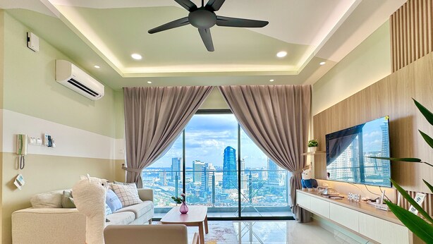 A modern living room with a sofa, a coffee table, and a large TV. There is a city view through the windows, and the room has a ceiling fan and recessed lighting.