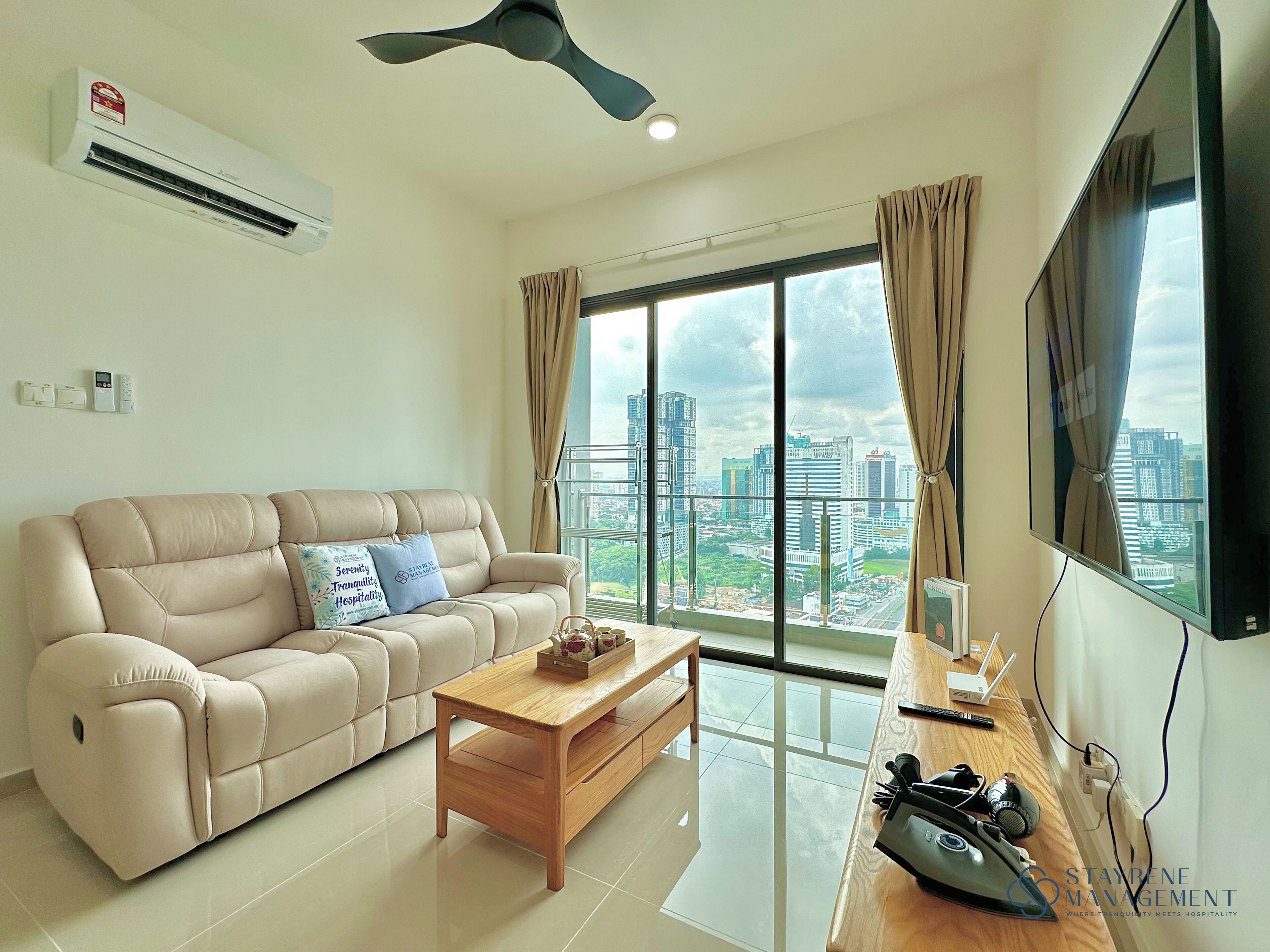 A modern living room with a beige sofa, a wooden coffee table, a ceiling fan, and a large window offering a city view.