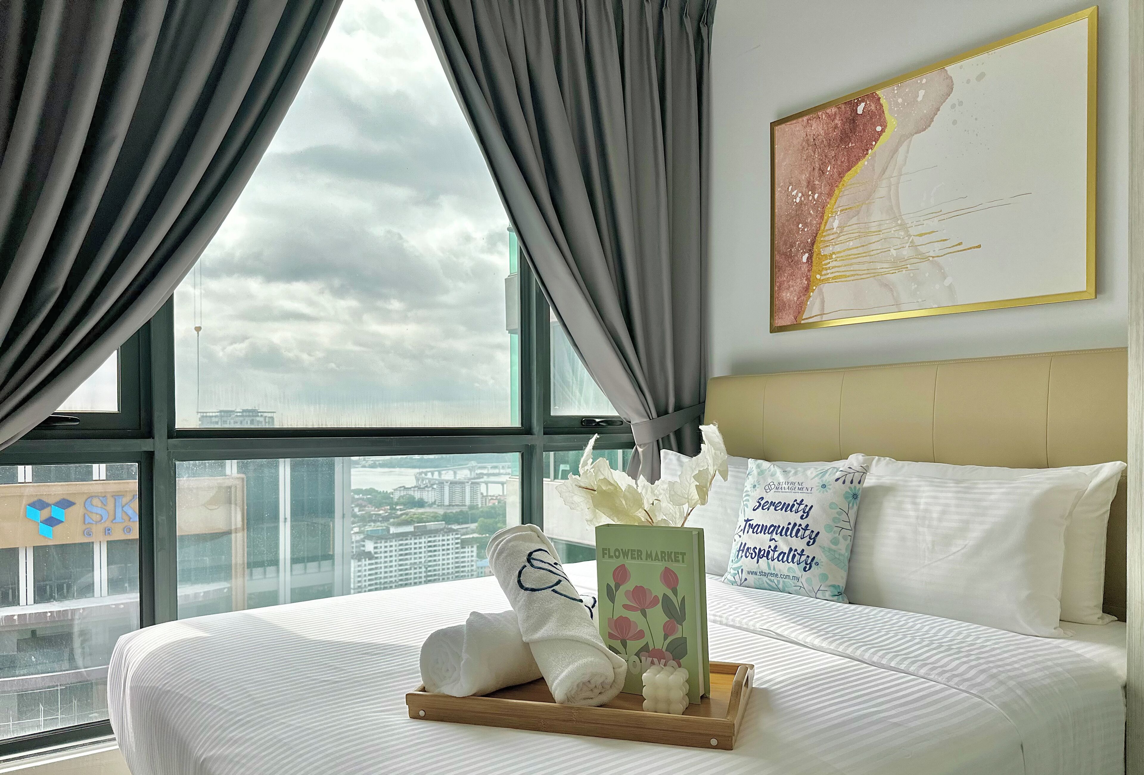 A hotel room with a bed, a tray with towels and a card, a window with curtains, and a wall with a framed picture.