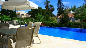 A poolside area with a table and chairs, a white umbrella, and a view of the pool and surrounding greenery. - Seaview 4 Bedroom villa with pool (Paphos)