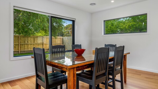 A dining area with a glass table and six chairs, large windows with a view of a wooden fence and greenery, and a red bowl on the table. - Ocean Oasis on Kenneth (Inverloch)