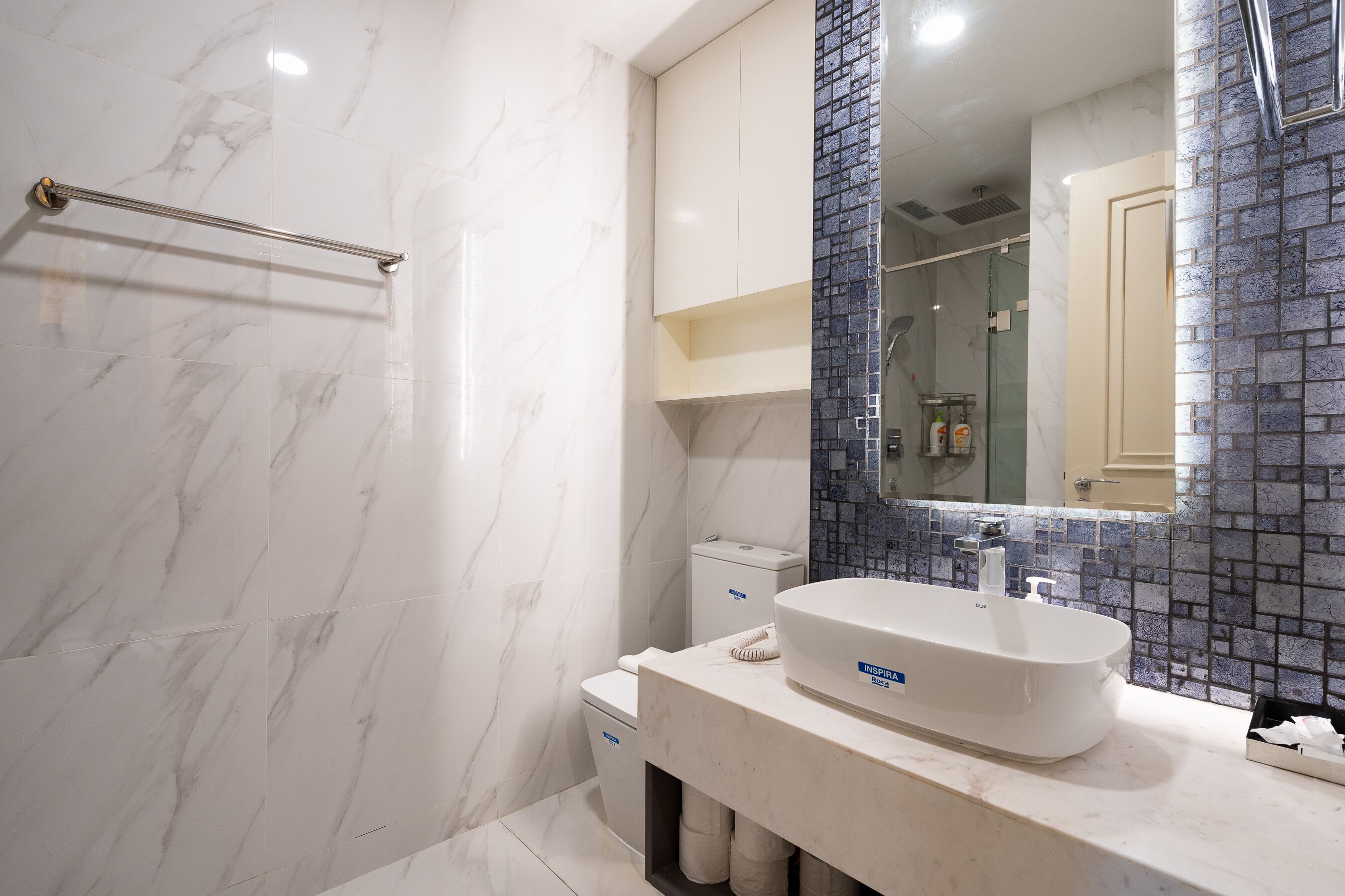 A bathroom with a white marble floor, a white sink with a soap dispenser, a glass shower enclosure, and a blue-tiled shower wall.