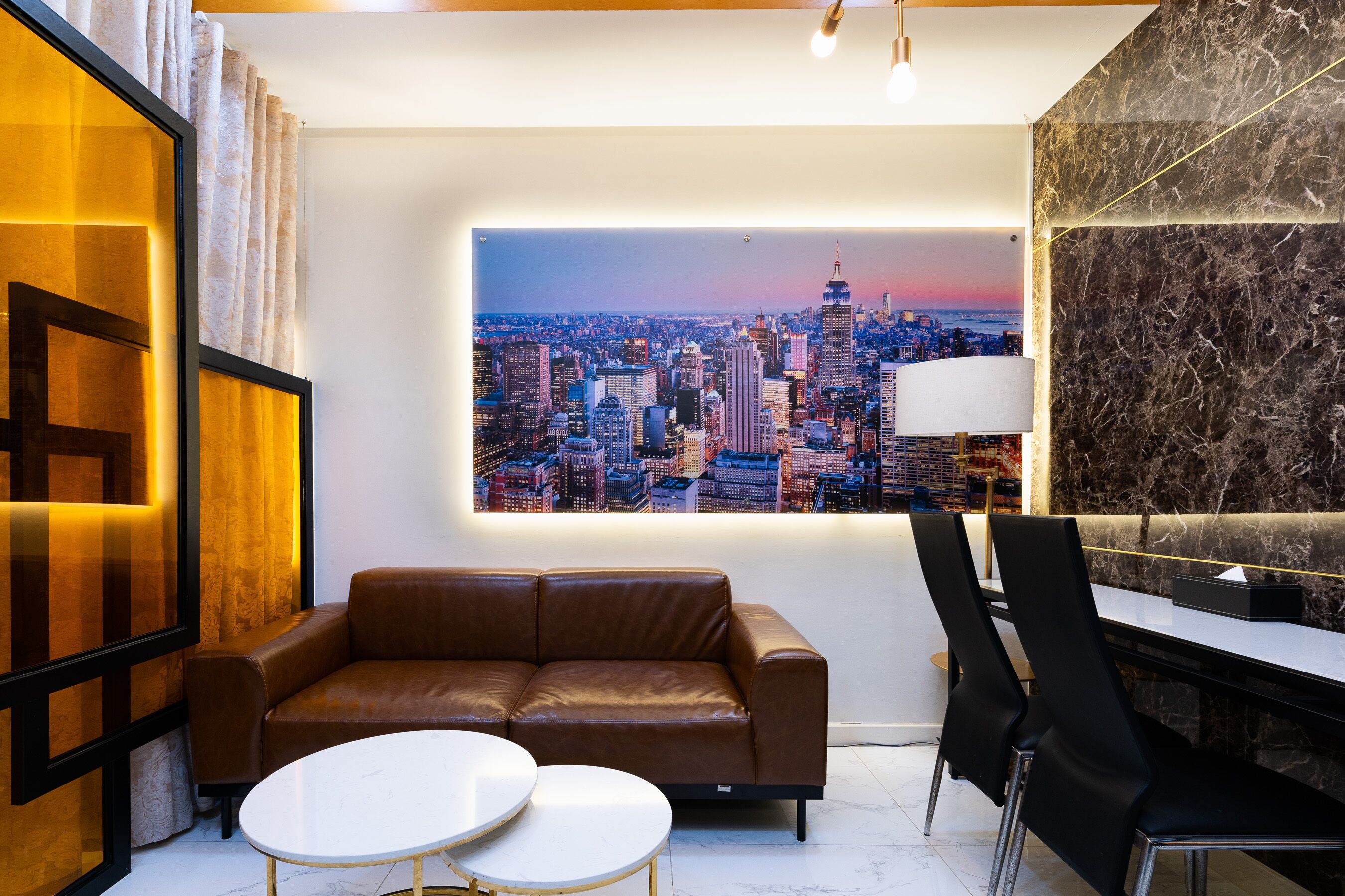 A modern living room with a cityscape view, a brown leather sofa, a white table, and black chairs.