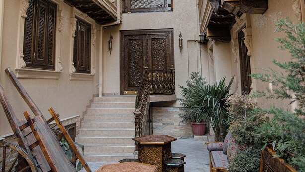A narrow courtyard with ornate wooden furniture, a staircase, and a potted plant.