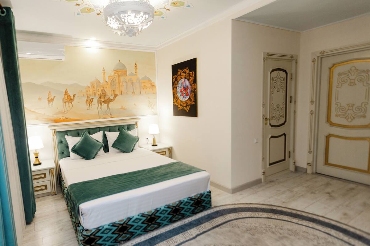 A bedroom with a bed, a nightstand, a lamp, a wall painting, a door, and a chandelier.