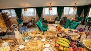 A buffet spread with various foods and drinks, green upholstered chairs, and a dining table in a well-lit room with large windows.