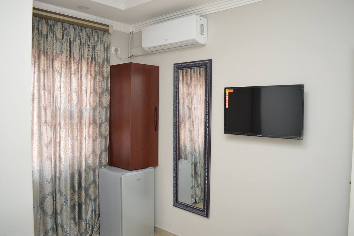 Una habitación con una televisión de pantalla plana en la pared, un armario de madera, un refrigerador y una cortina con estampado floral.