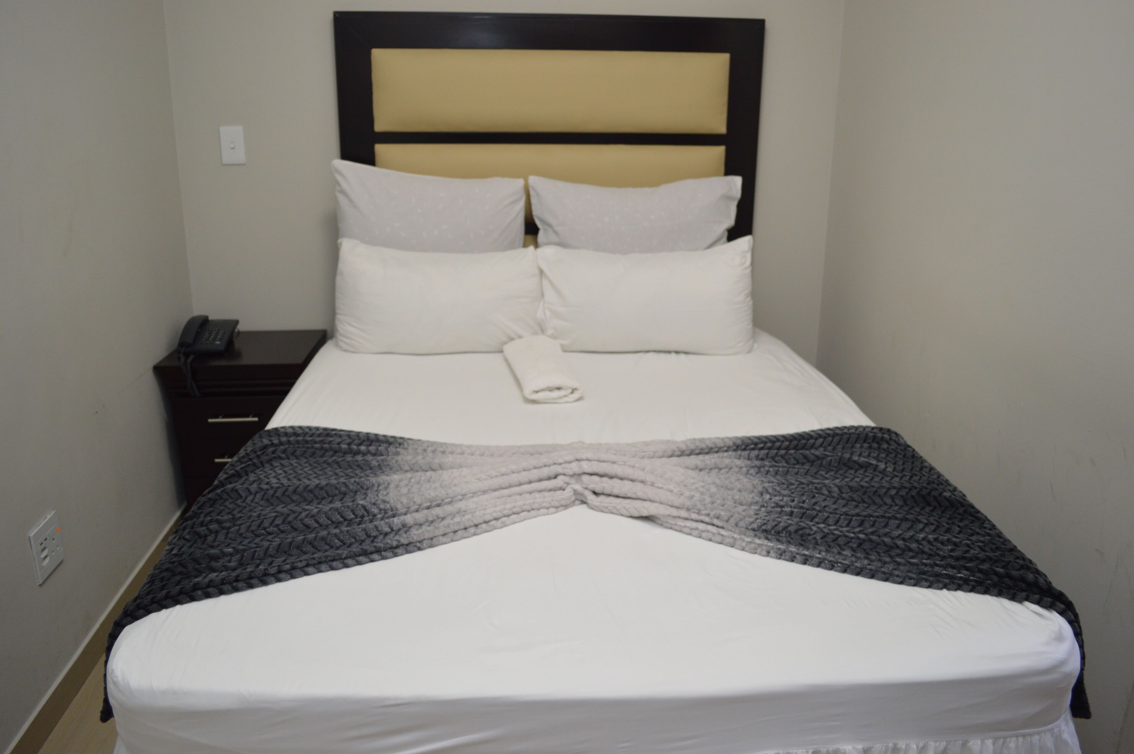 A neatly made bed with white linens and a folded towel, a dark wooden nightstand with a phone, and a headboard with a beige panel.