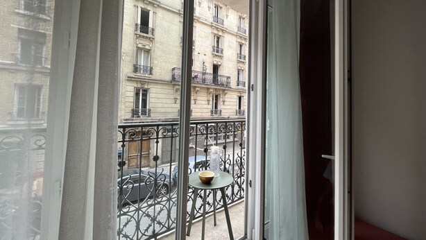 A room with a large window overlooking a street with cars and a building with balconies. - Bel Appartement Haussmannien à Montparnasse (Paris)