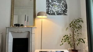 A room with a white fireplace, a gold-framed mirror, a floor lamp, a black and white abstract painting, and a potted plant. - Bel Appartement Haussmannien à Montparnasse (Paris)
