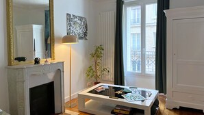 A living room with a white fireplace, a large mirror, a floor lamp, a coffee table with a Scrabble game, and a view of the outside balcony through a window. - Bel Appartement Haussmannien à Montparnasse (Paris)