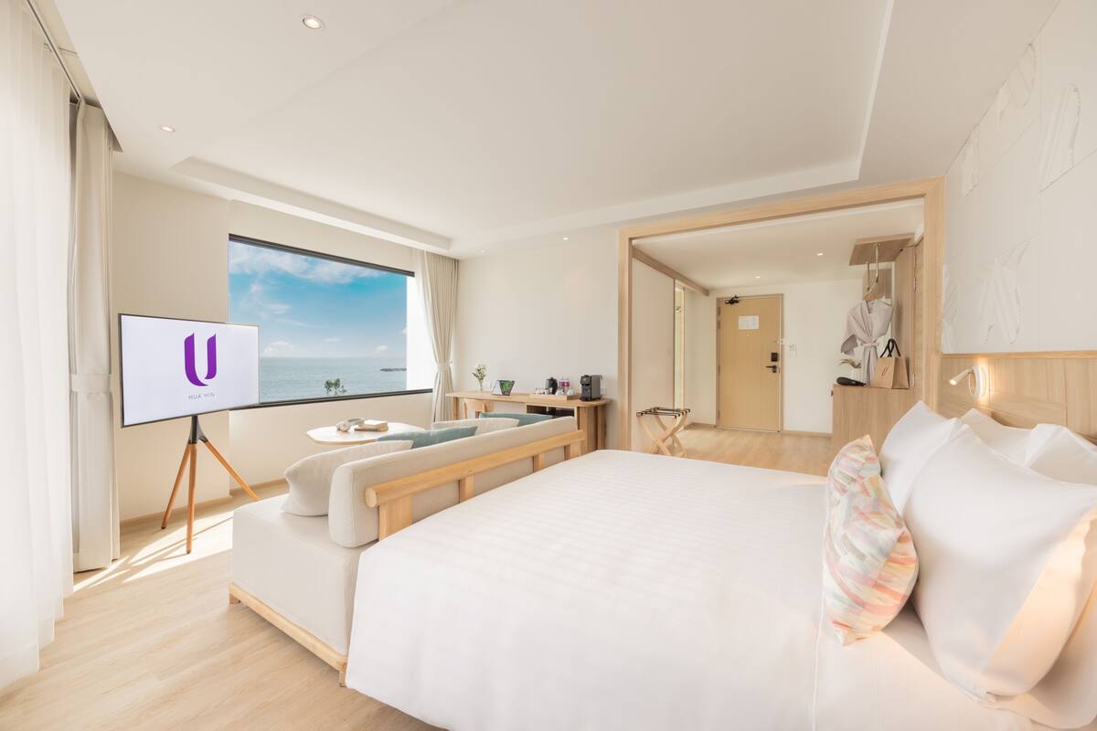 Une chambre d’hôtel moderne avec un grand lit, un téléviseur posé sur un meuble, une vue sur la mer, une petite table avec une plante et une table de chevet en bois.