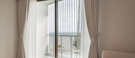 Habitación de hotel con un ventanal grande, una cama, una mesita con un libro y una cámara, y un balcón con vistas al mar.